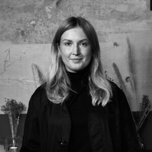 Porträt einer jungen Frau mit blondem Haar, die ruhig und selbstbewusst in die Kamera blickt, vor einer hellen, strukturierten Wand im minimalistischen Setting.